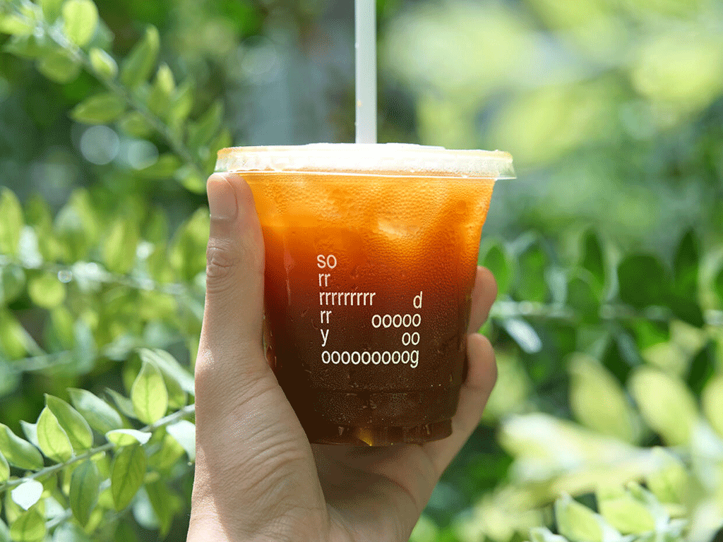 Disposable iced coffee cup – YamiPak Coffee PET cold brew cup – clear cup with flat lid and logo – handheld in green outdoor background, close-up angle