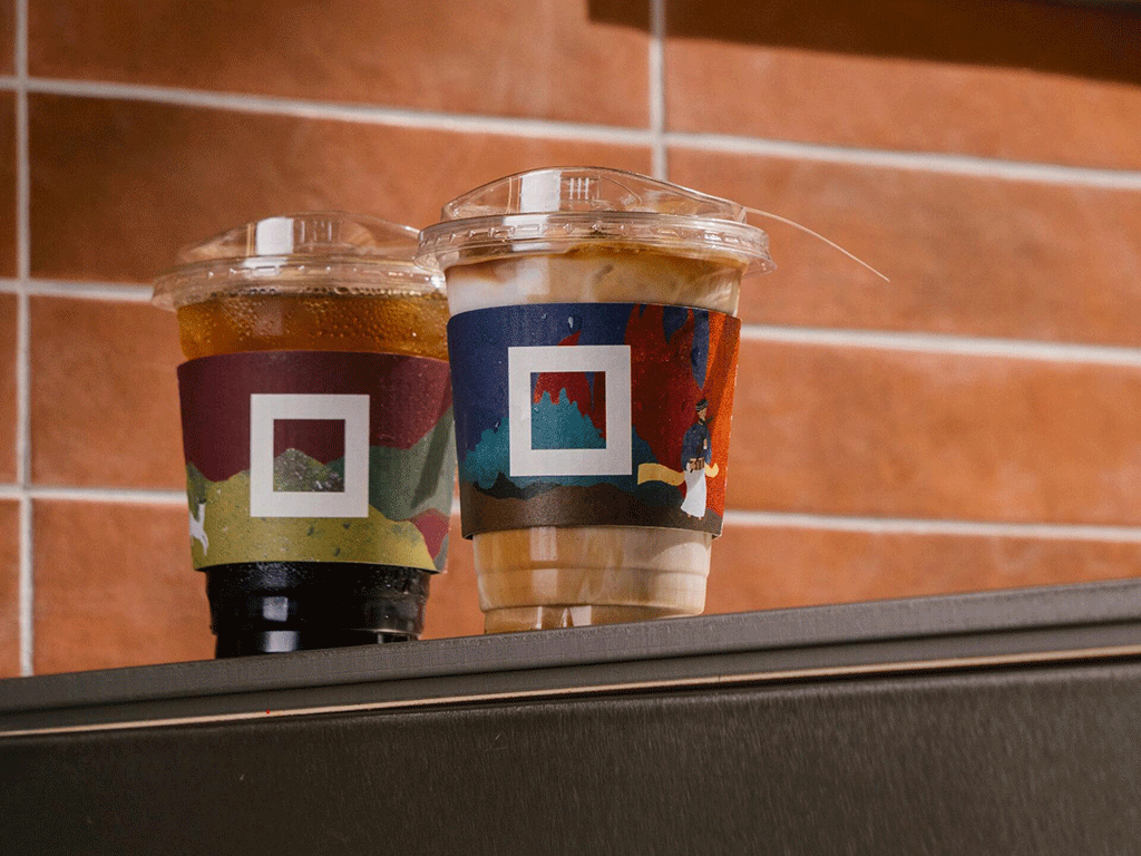Two disposable plastic cups with custom sleeves and clear lids, placed on a black surface against a tiled background.