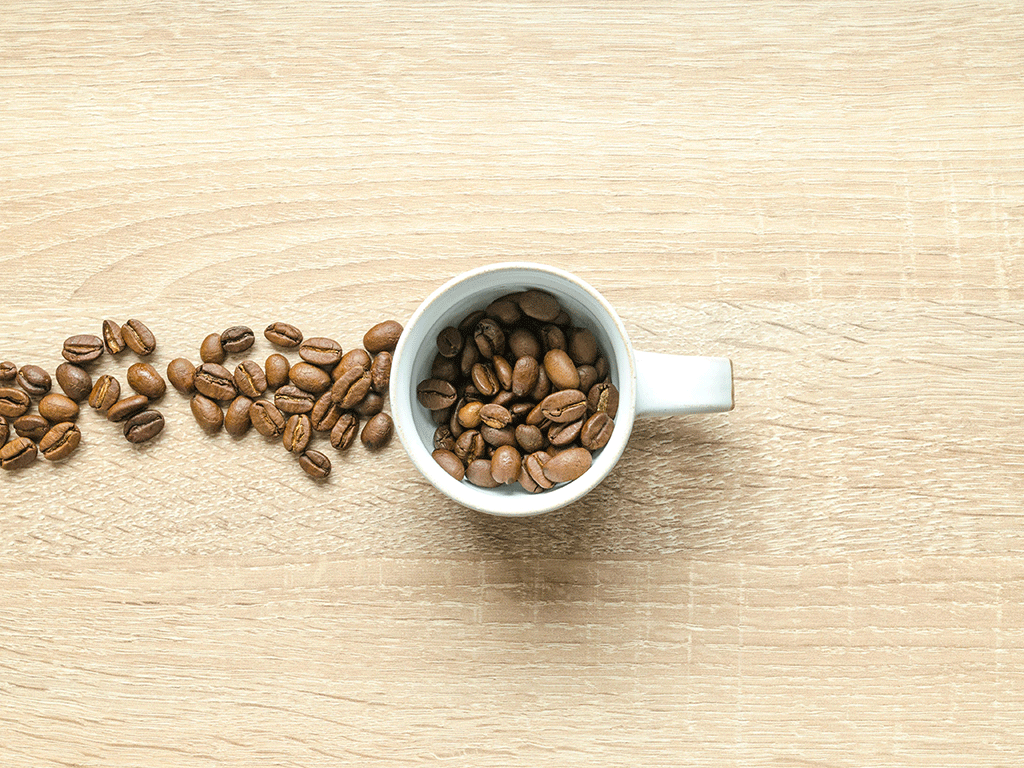 Coffee beans spilling from a cup on a light wooden surface, representing roasted beans ready for brewing and flavor preservation