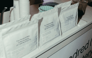 Three white coffee bags with a rough matte finish, featuring a minimalist design with bold text and coordinates on the front.