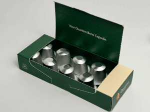 An open coffee capsule box in dark green revealing ten aluminium capsules inside, with the text "Your Quarters Brew Capsule" printed on the inner lid.