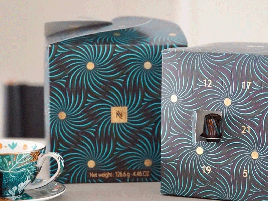 Two coffee capsule gift boxes with a dark teal and purple swirl pattern and gold dot details, displayed alongside a decorative espresso cup and saucer.