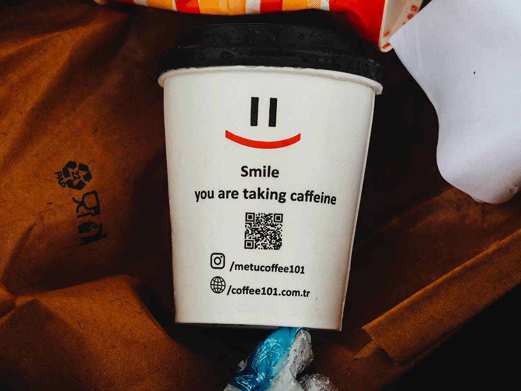 A single-use paper coffee cup with a printed QR code and social media handles resting in a recycling bin.